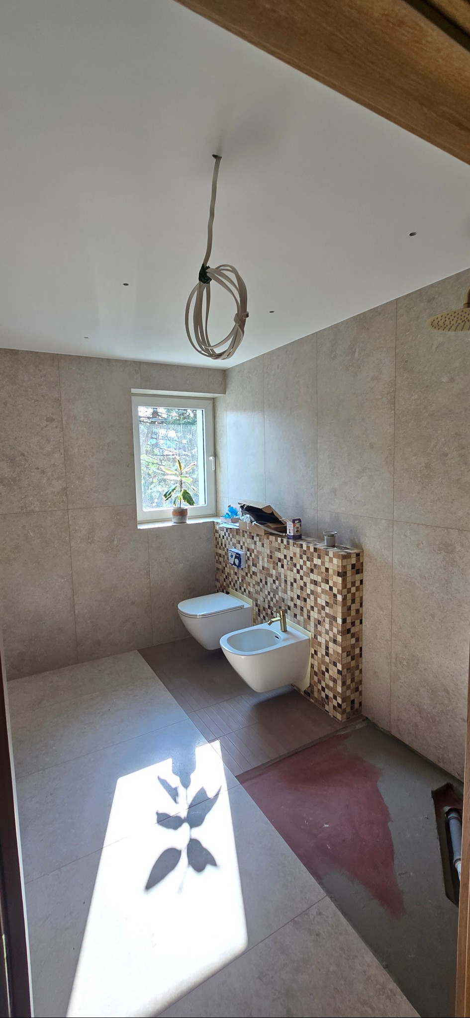 Jasna łazienka w trakcie remontu: beżowe płytki, mozaika za toaletą i bidetem, widoczne kable, cień rośliny na podłodze. Etap prac wykończeniowych.