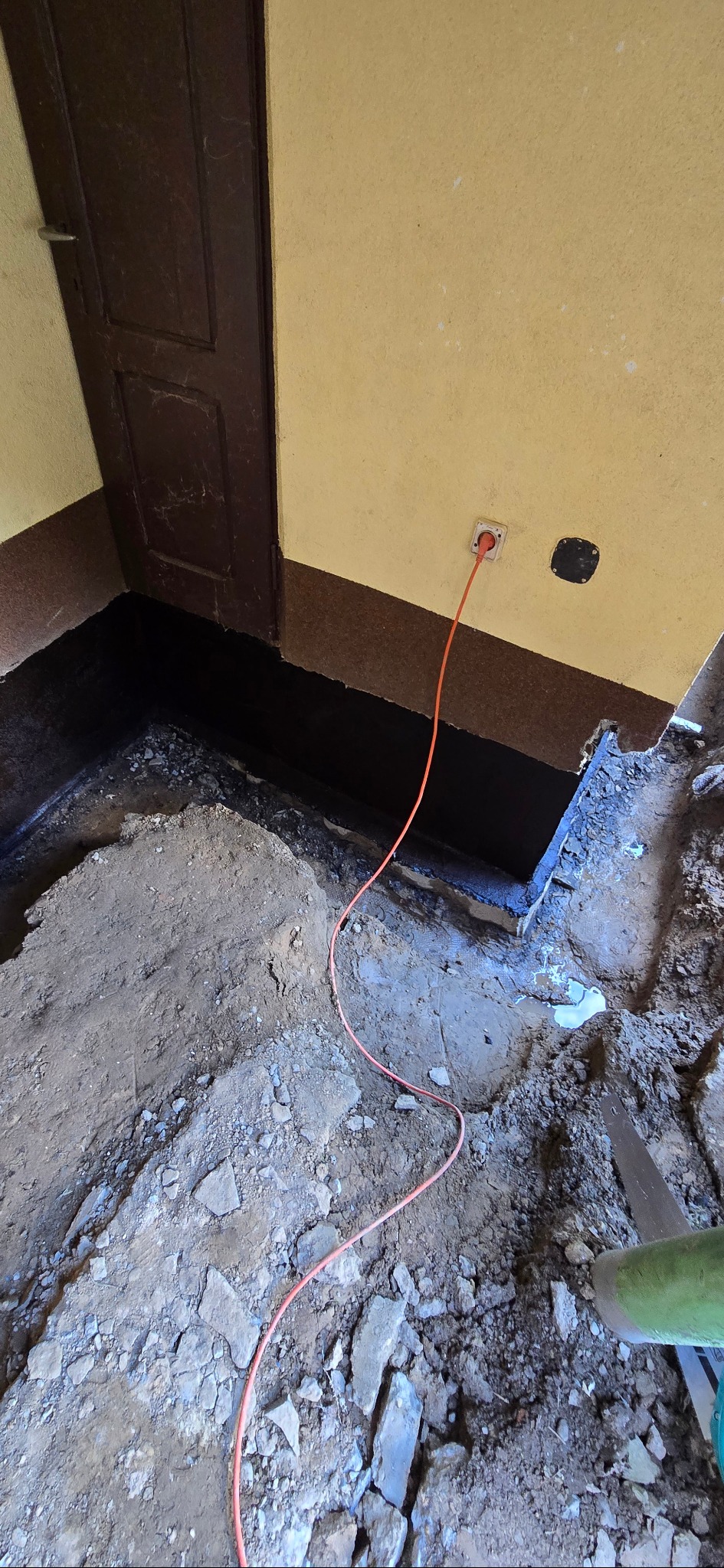 Przygotowanie do wylewki: wykopana podłoga, zabezpieczone ściany, wystający kabel zasilający. Widoczny fragment drzwi i ściany w kolorze żółtym.