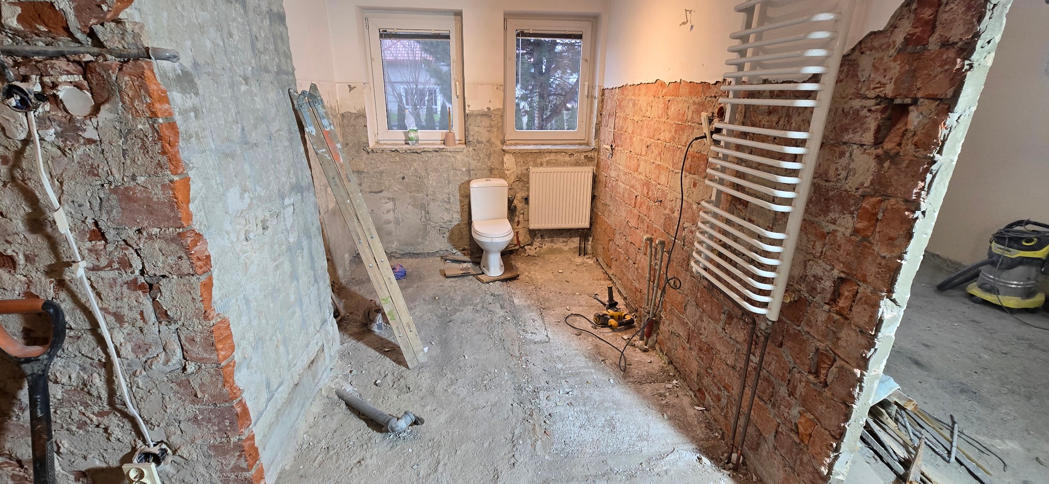 Łazienka w trakcie remontu: odsłonięta cegła, nowa toaleta, grzejnik, drabina i narzędzia. Widoczne ślady prac budowlanych na ścianach i podłodze.