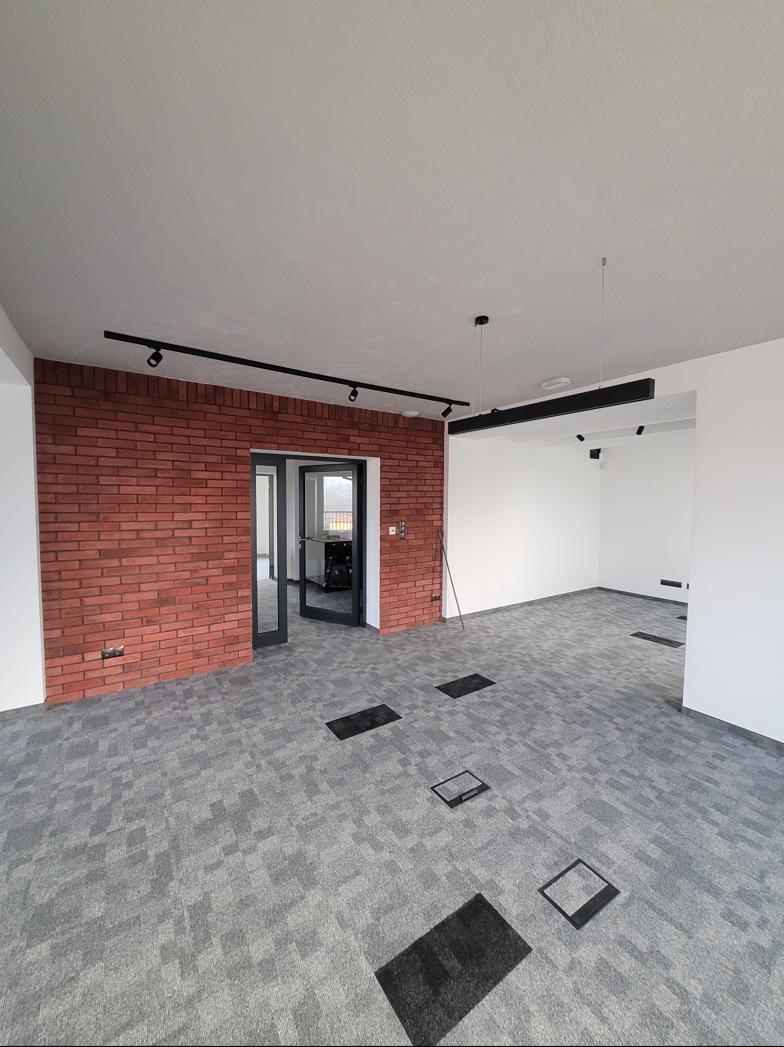 Surowe wykończenie biura: ceglana ściana, szara wykładzina dywanowa, czarne lampy sufitowe. Minimalizm i nowoczesność w przestrzeni biurowej.