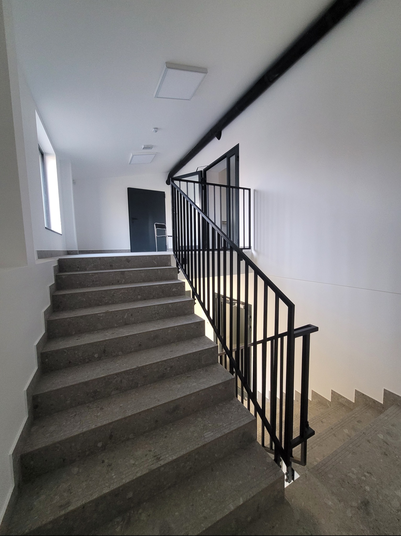 Klatka schodowa z szarymi stopniami i czarną metalową balustradą. Białe ściany, sufit z oświetleniem. Nowoczesny design.
