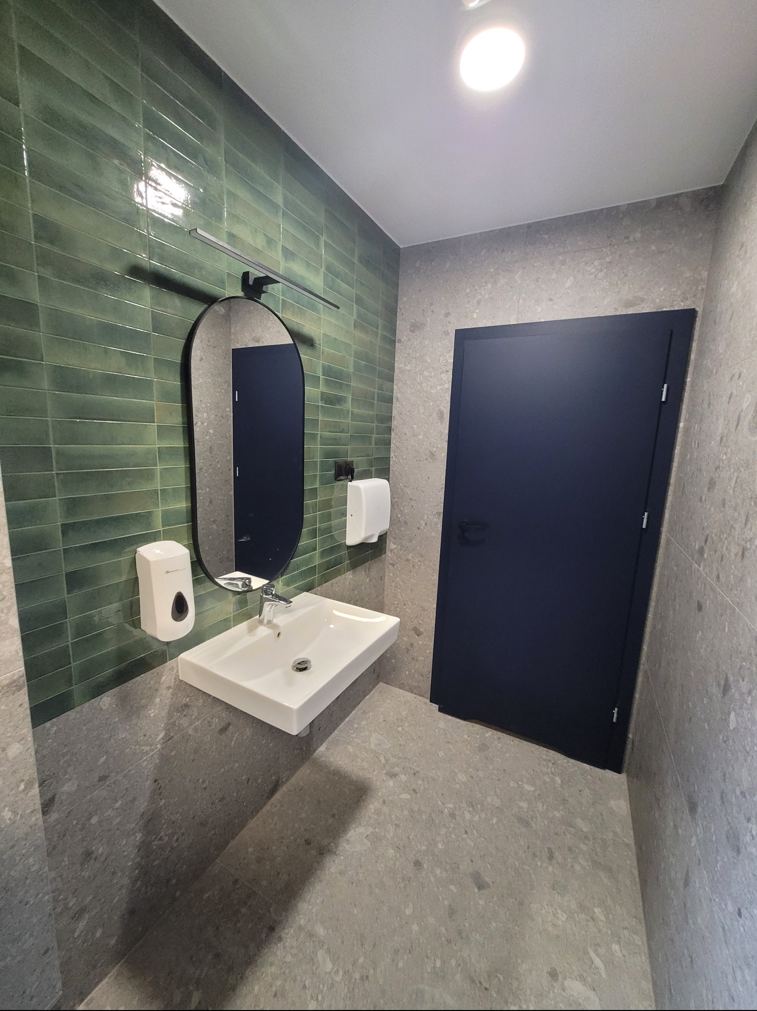 Nowoczesna łazienka z umywalką nablatową, zielonymi płytkami na ścianie i owalnym lustrem. Minimalistyczny design, szare płytki na podłodze i ścianach.