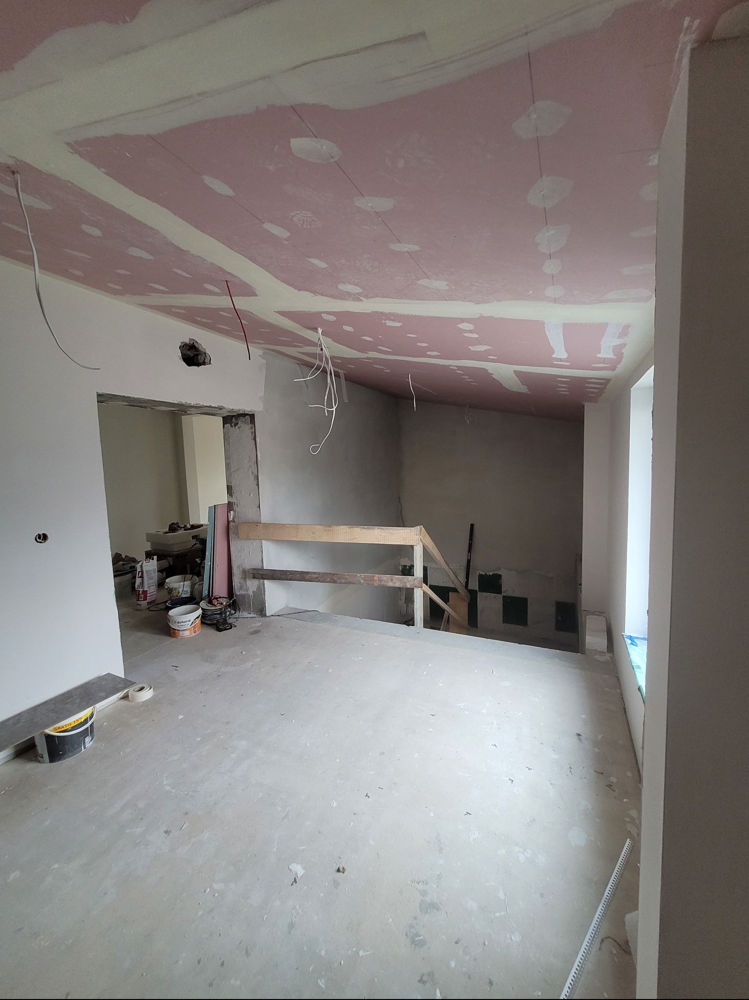 Wnętrze w trakcie prac wykończeniowych: sufit z płyt gipsowo-kartonowych w kolorze różowym, ściany w stanie surowym, widoczne kable i narzędzia. Pomieszczenie w trakcie remontu.