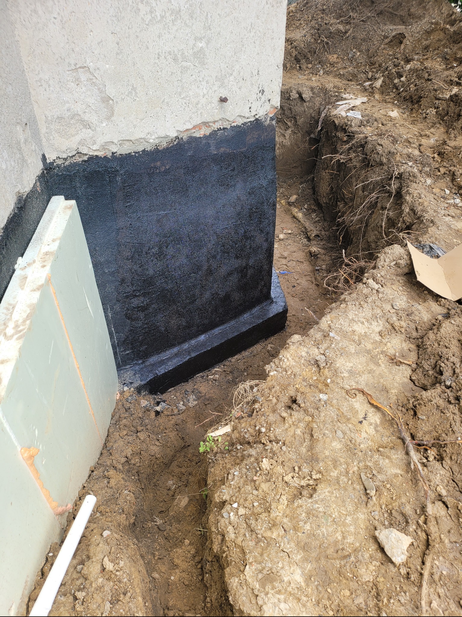 Hydroizolacja fundamentu budynku w trakcie prac ziemnych, widoczna czarna powłoka izolacyjna i płyta styropianowa. Wykop wokół fundamentu.