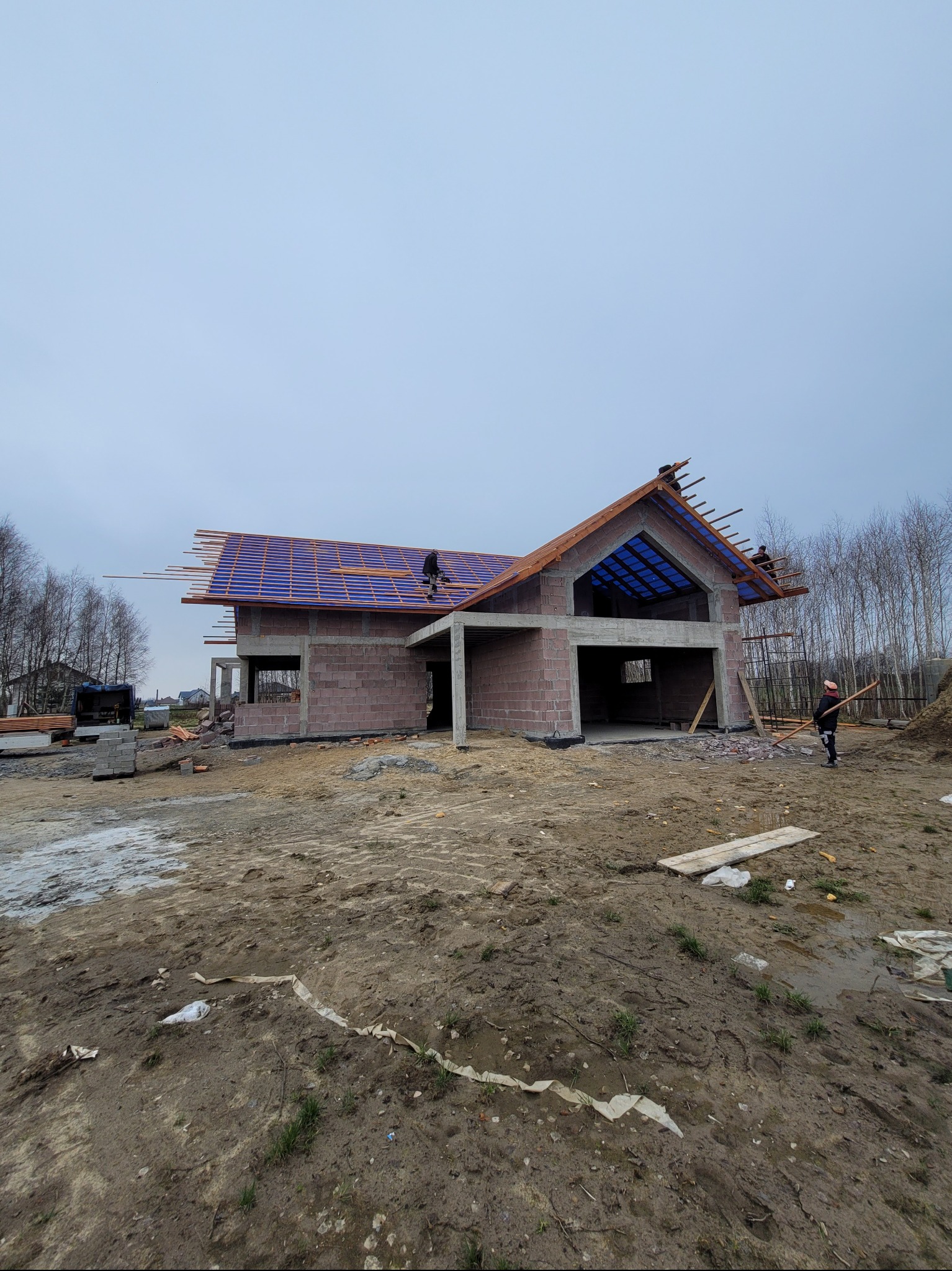 Budowa domu jednorodzinnego w stanie surowym otwartym. Widoczni pracownicy montujący konstrukcję dachu, niebieska folia dachowa i ceglany budynek.