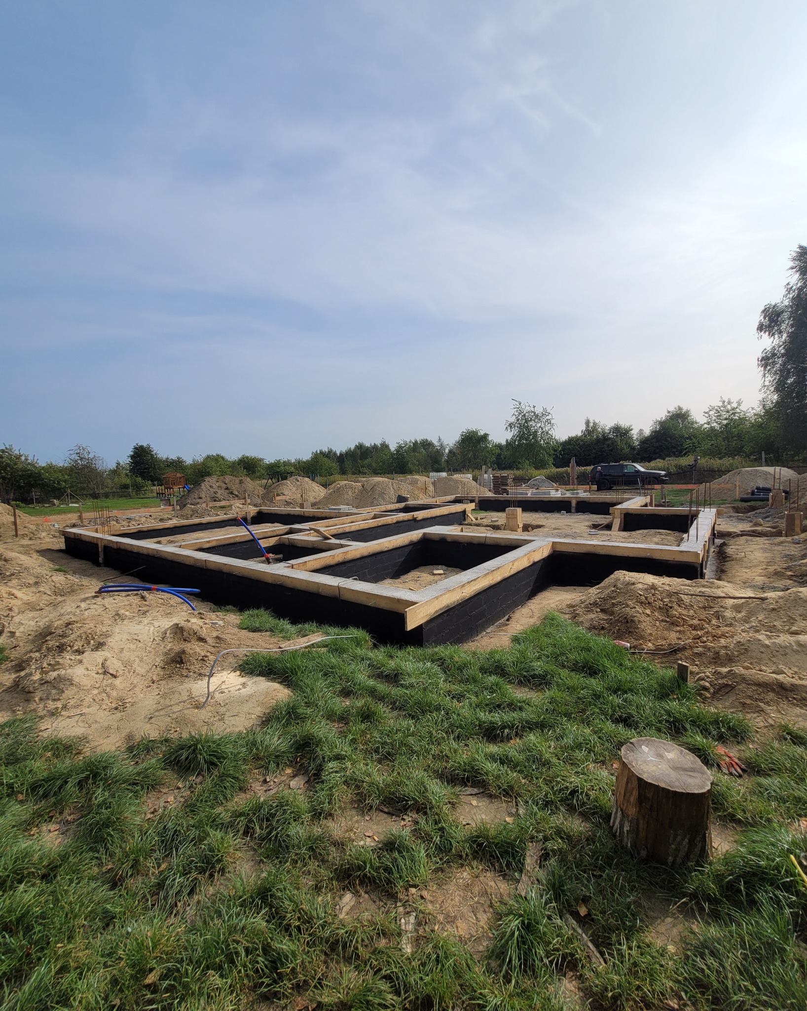 Fundamenty domu w trakcie budowy, widoczne drewniane szalunki i izolacja przeciwwilgociowa. Świeża trawa w pierwszym planie, w tle zieleń i błękitne niebo.