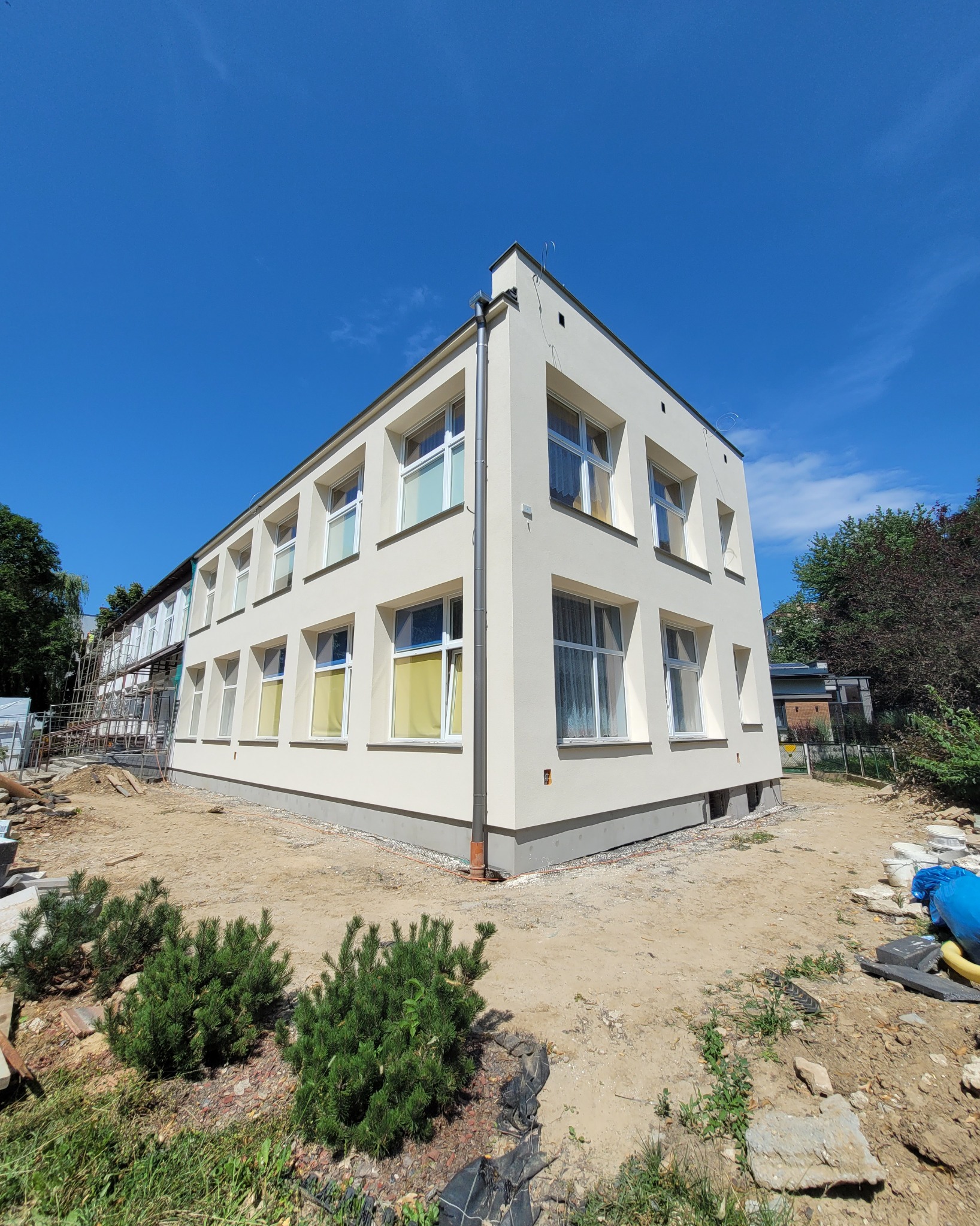 Odnowiona elewacja budynku z nowymi oknami, widziana z perspektywy ziemi w słoneczny dzień. Częściowo widoczne rusztowania i elementy krajobrazu.