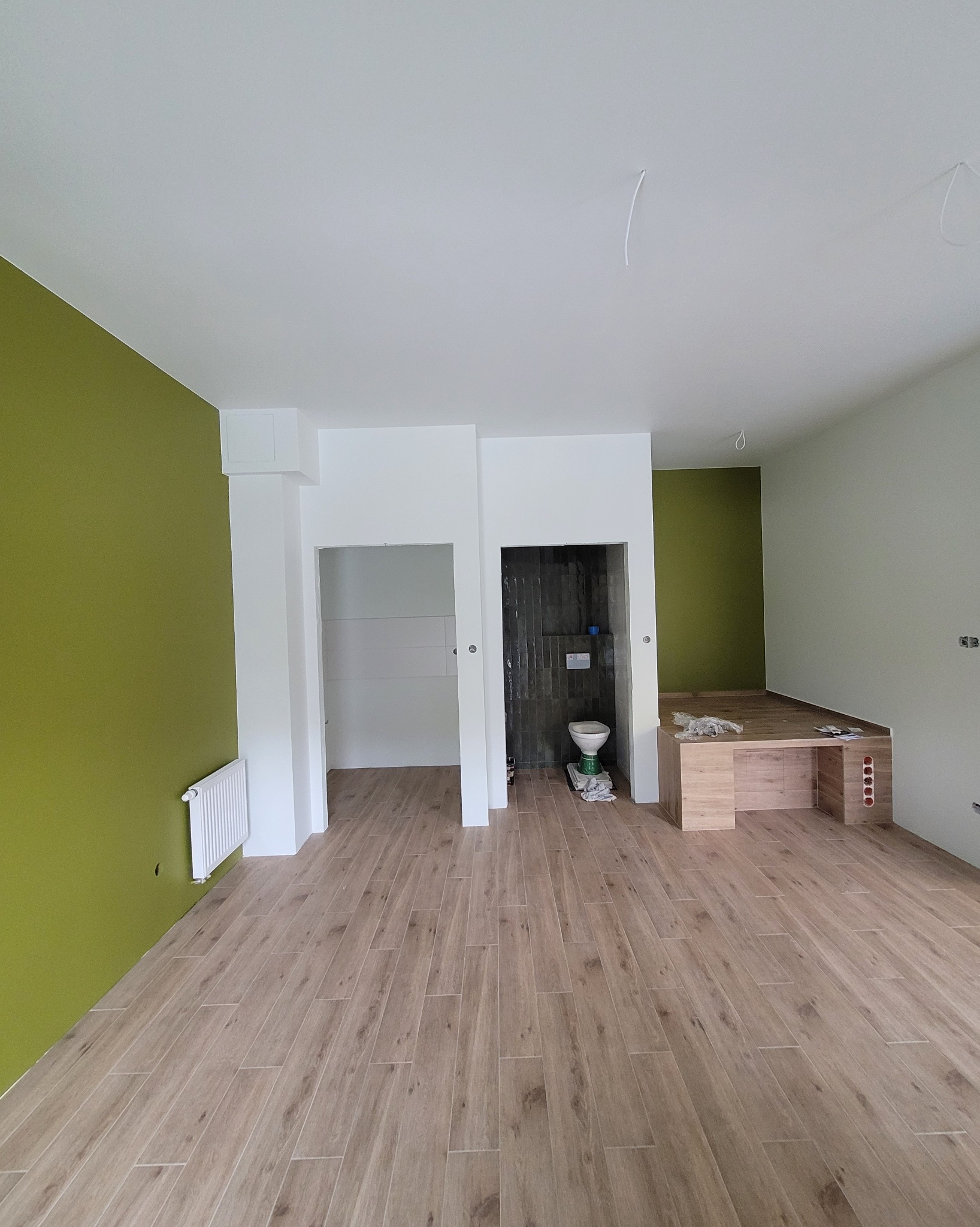 Wnętrze po remoncie: drewnopodobna podłoga, ściana w odcieniach zieleni, wykończona łazienka z toaletą i fragment zabudowy z drewnianymi elementami.