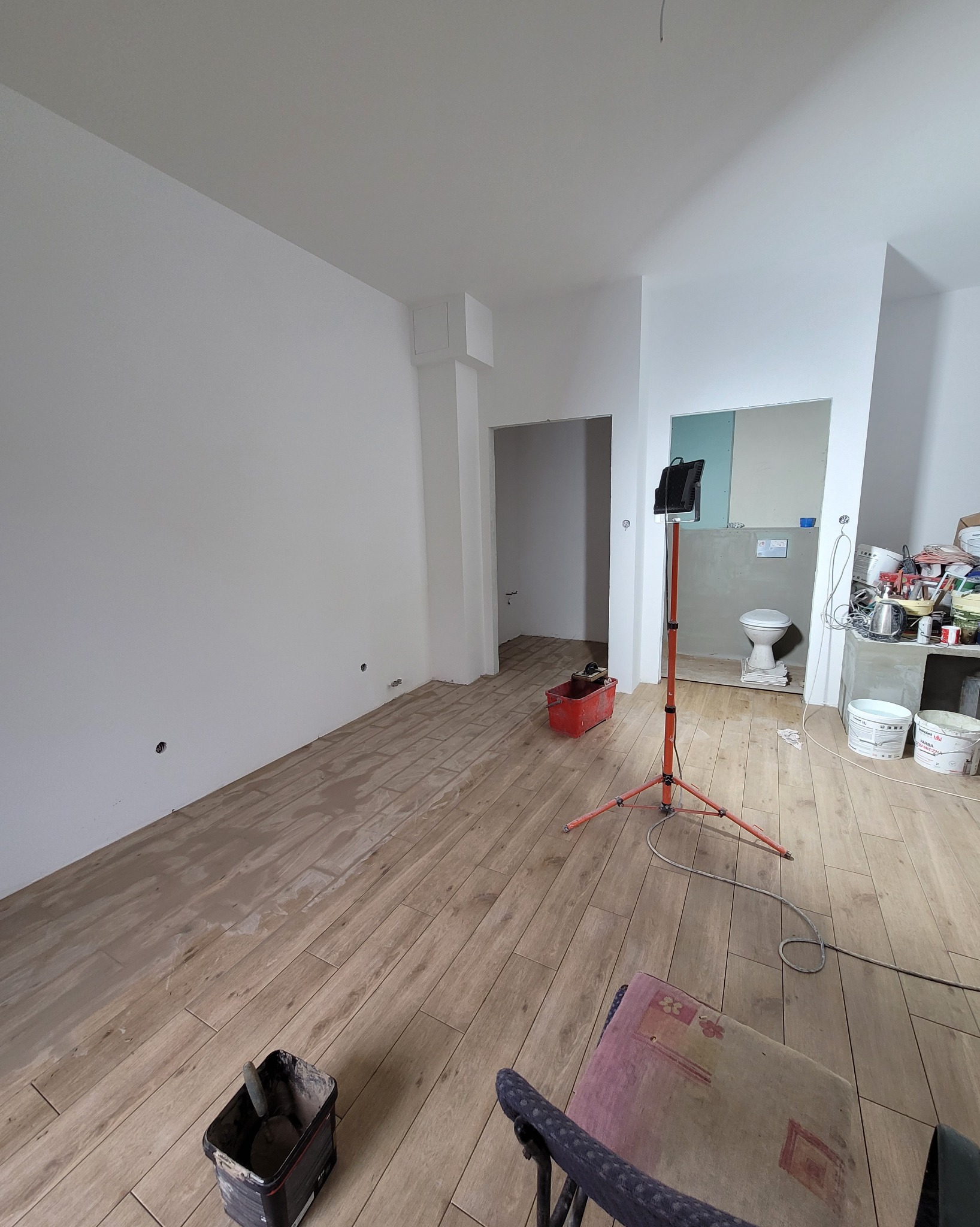 Wnętrze w trakcie remontu: podłoga wyłożona płytkami imitującymi drewno, ściany przygotowane do malowania, widoczne narzędzia i oświetlenie budowlane.