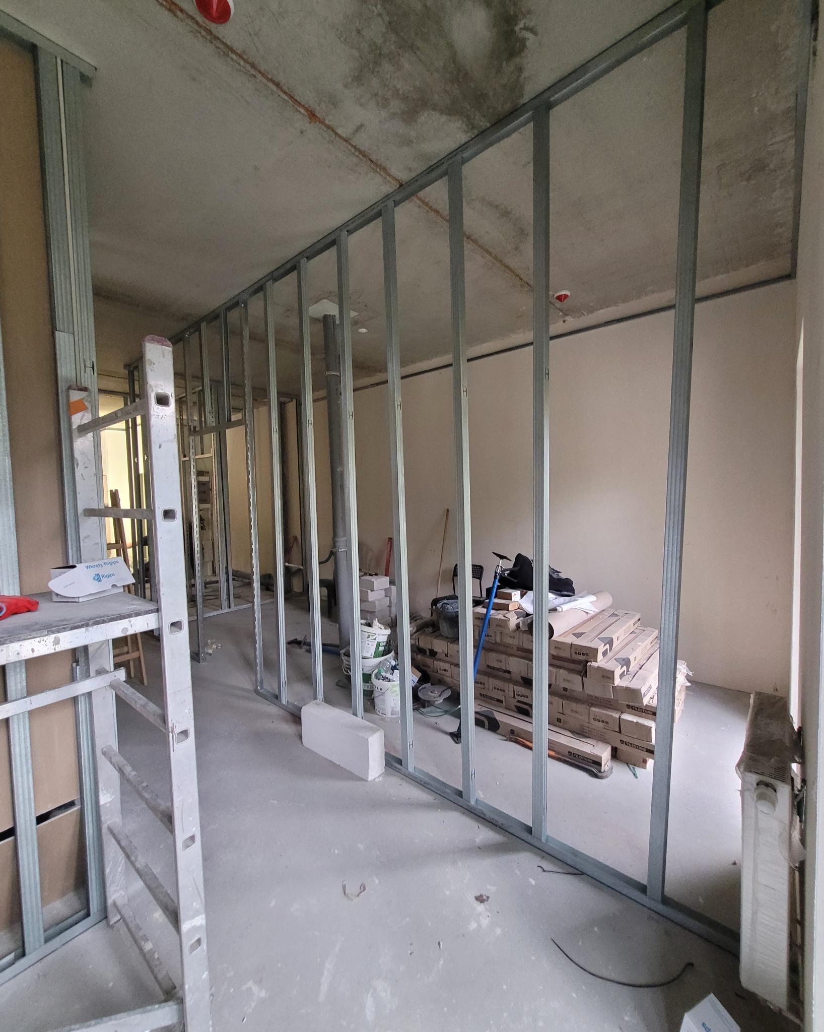 Montaż metalowego szkieletu ścian działowych w surowym wnętrzu. Widoczna drabina, narzędzia i materiały budowlane. Stan surowy, przygotowanie do dalszych prac wykończeniowych.