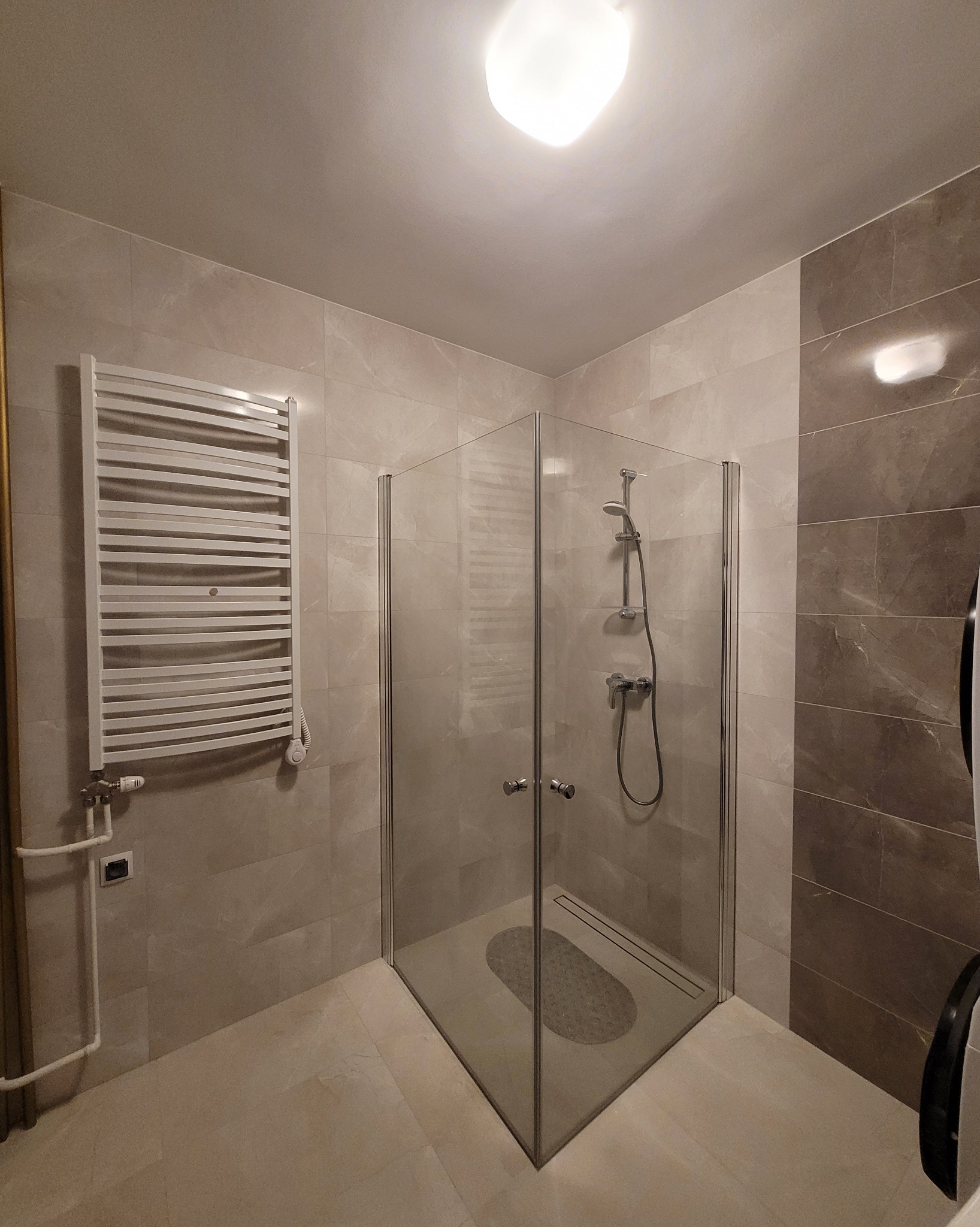 Nowoczesna łazienka z kabiną prysznicową typu walk-in, grzejnikiem drabinkowym i beżowymi płytkami imitującymi marmur. Minimalistyczny design, stonowana kolorystyka.