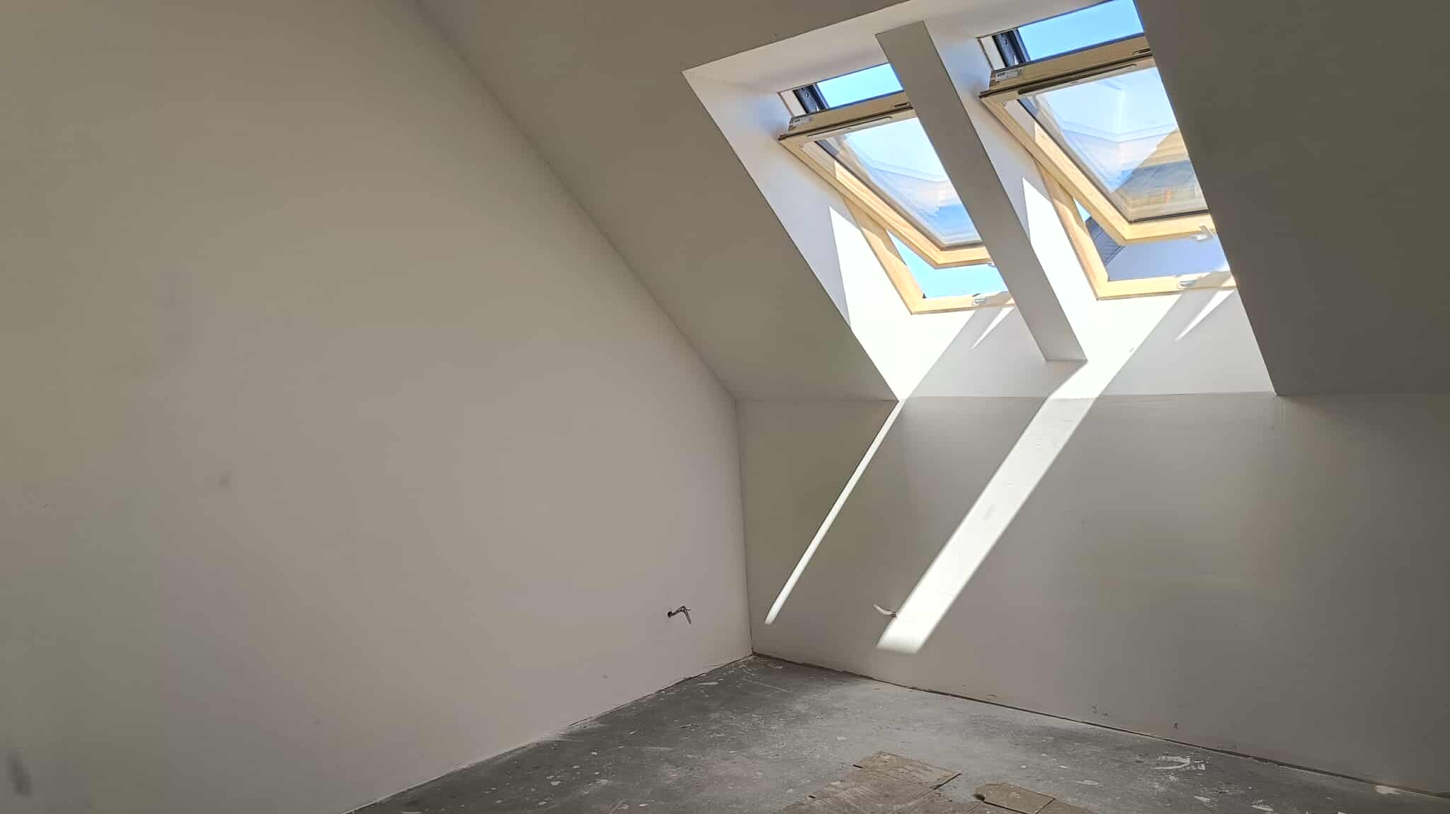 Poddasze po malowaniu na biało, z dwoma oknami dachowymi w drewnianych ramach, wpadające promienie słoneczne, szary beton na podłodze, widok z dołu.