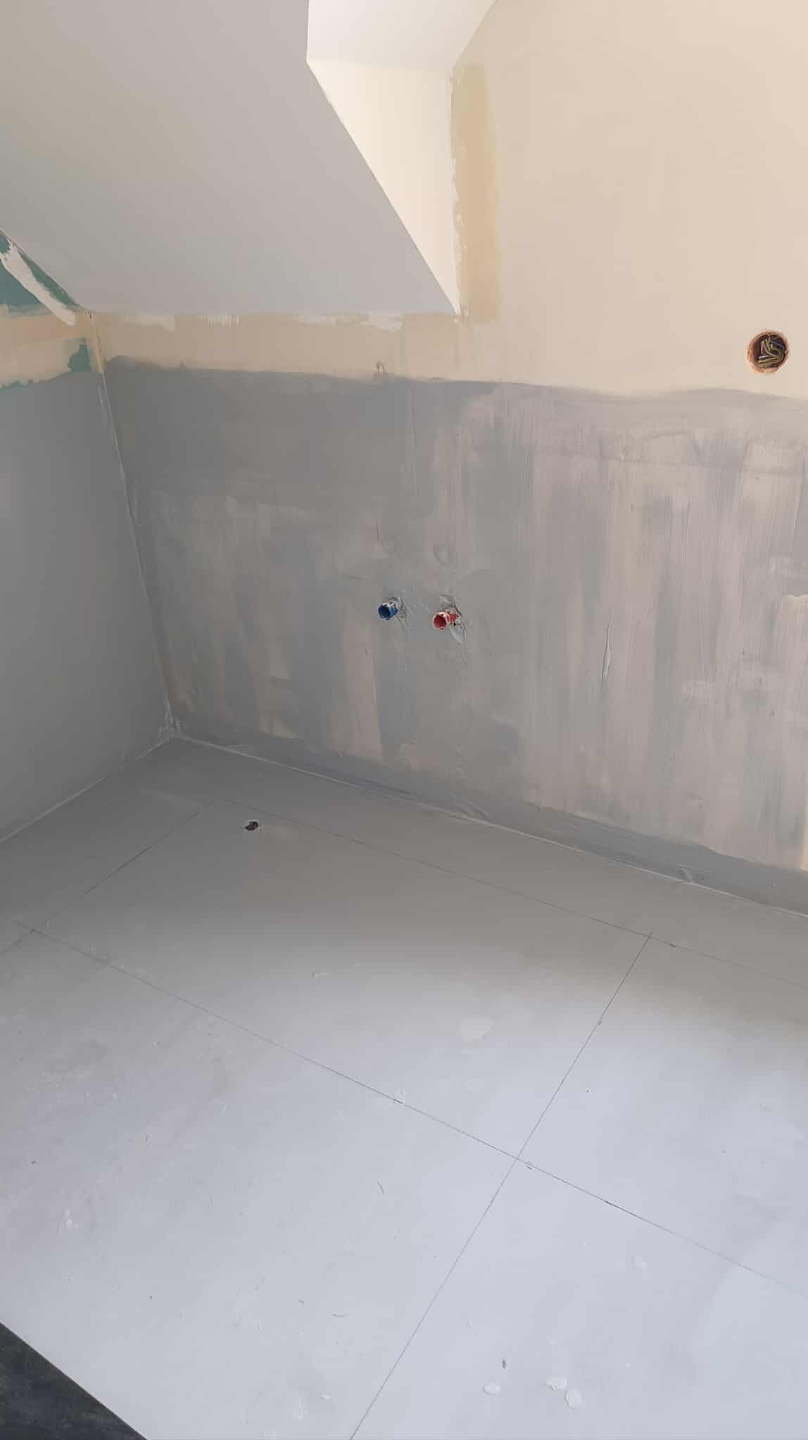 Wnętrze łazienki w trakcie remontu: szare ściany po gruntowaniu, białe płytki na podłodze, wystające rury instalacji wodnej. Widoczne ślady po szpachlowaniu.