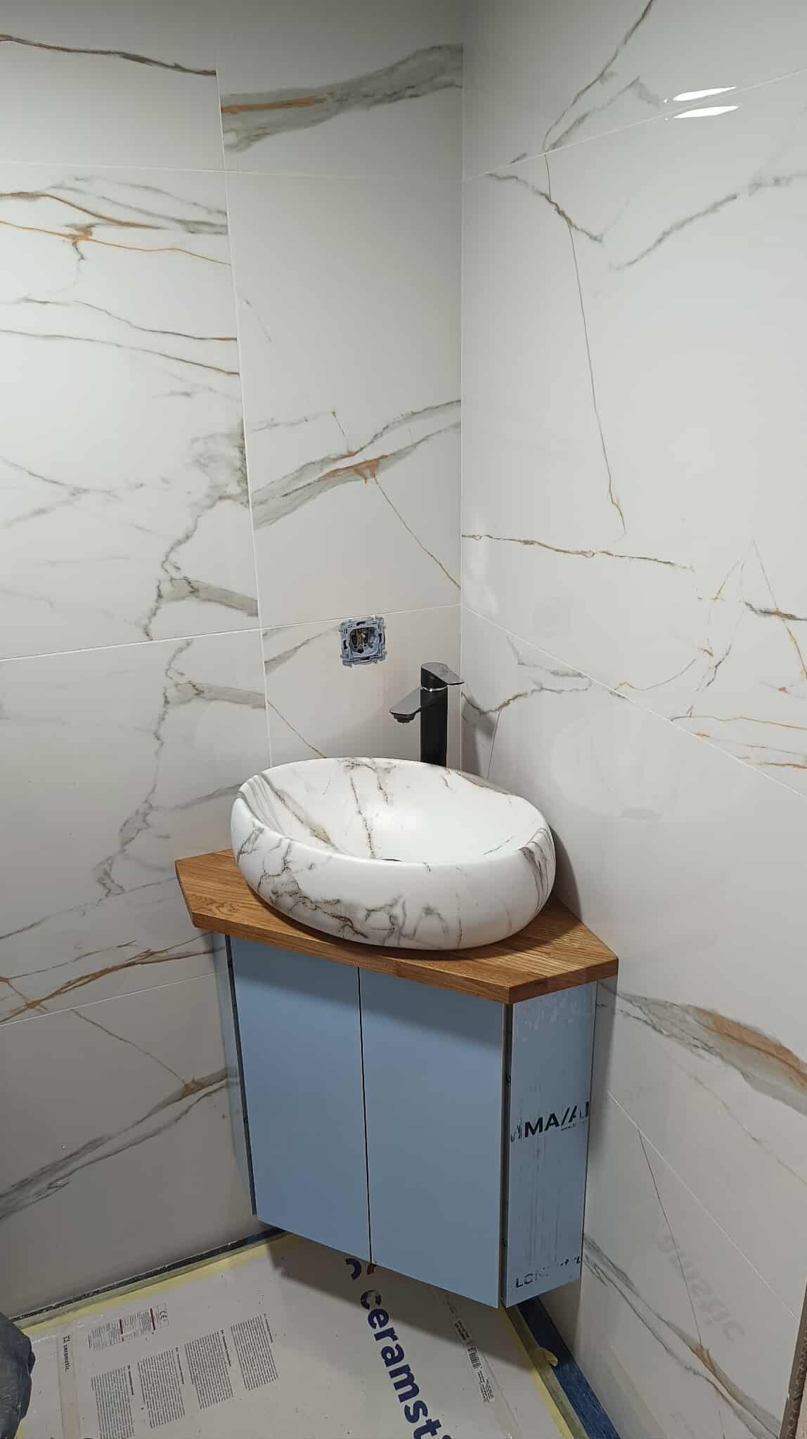 Wykończona łazienka: marmurowe płytki, owalna umywalka na drewnianym blacie i niebieska szafka. Widoczna instalacja elektryczna. Nowoczesny design.
