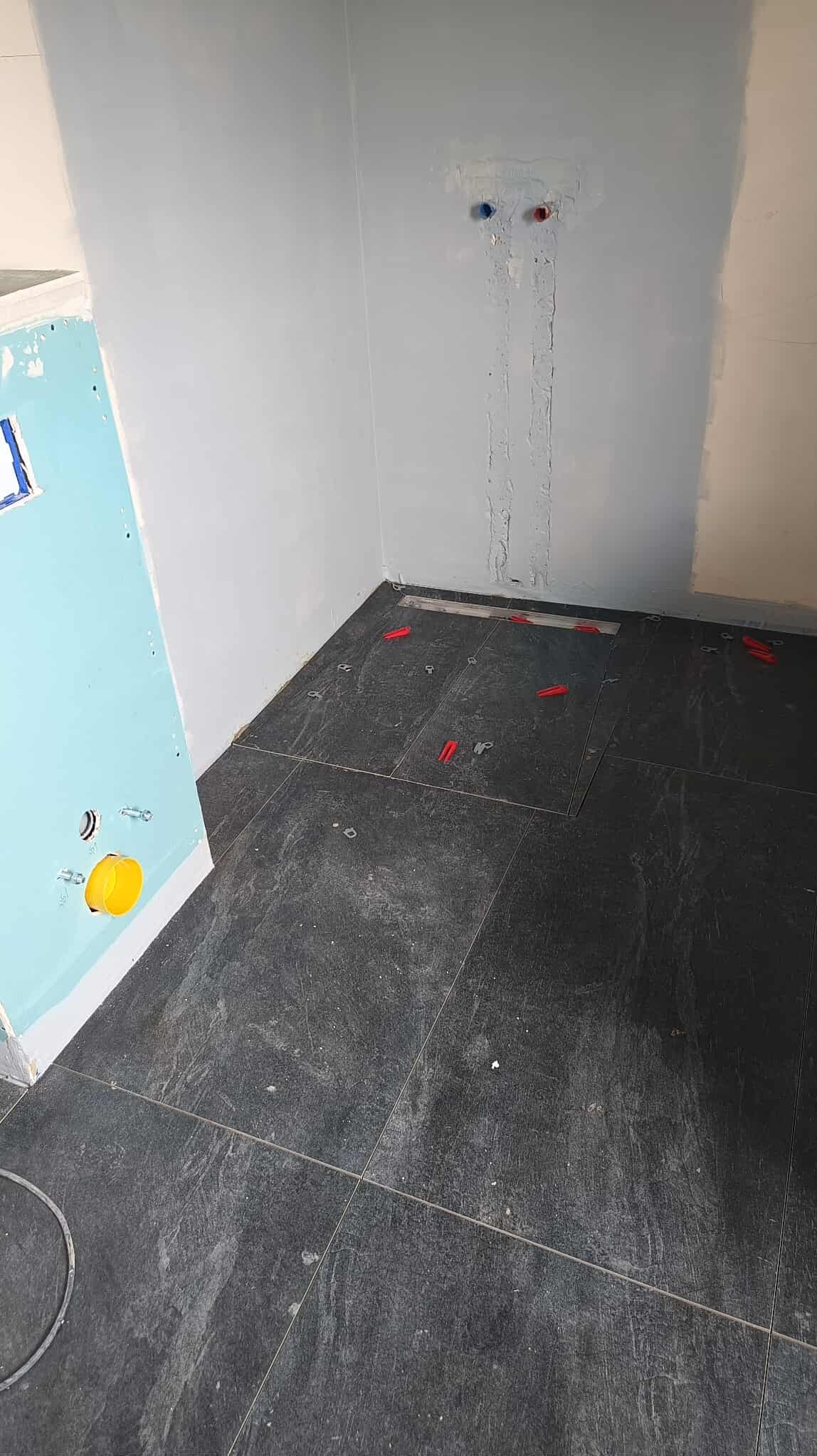 Ciemna, grafitowa podłoga z płytek w trakcie układania w łazience. Widoczne czerwone kliny poziomujące, szara ściana i fragment niebieskiej płyty gipsowej.