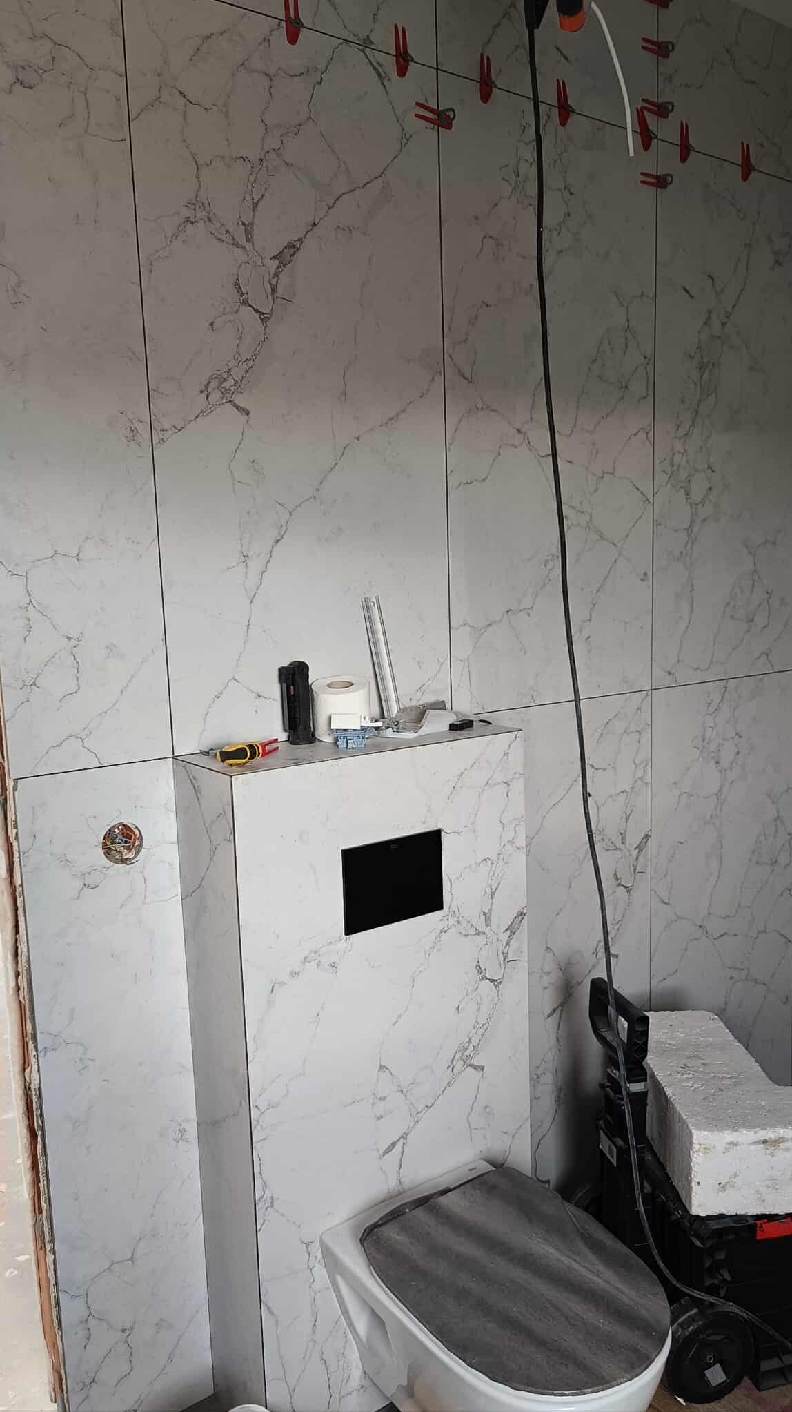 Wykończona toaleta z marmurowymi płytkami, widoczne narzędzia glazurnicze i poziomica. Czerwone klipsy do poziomowania płytek na ścianie.