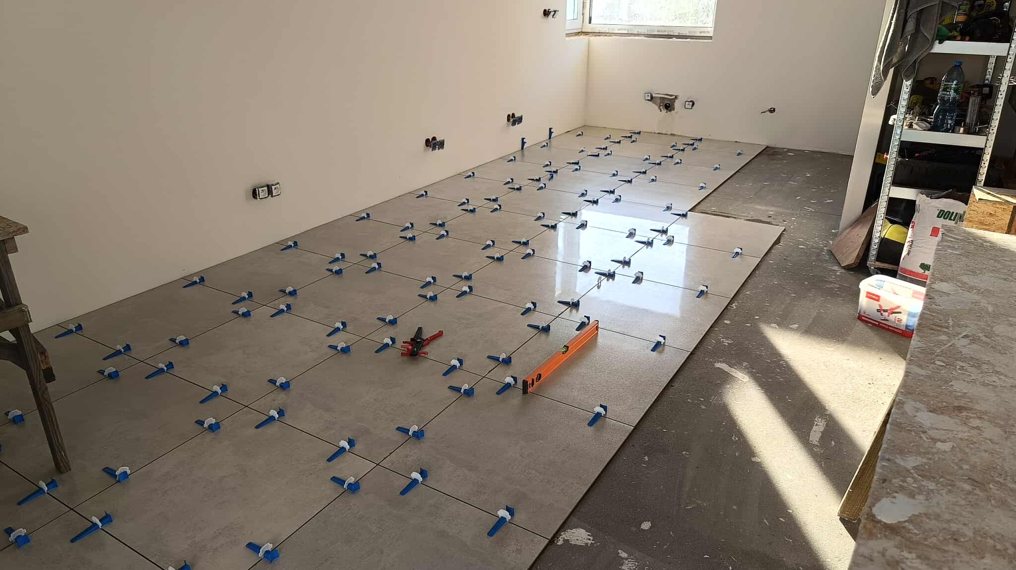 Układanie jasnych płytek podłogowych z użyciem systemu poziomowania. Widoczna poziomica i narzędzia glazurnicze. Częściowo wykończona podłoga w jasnym pomieszczeniu.