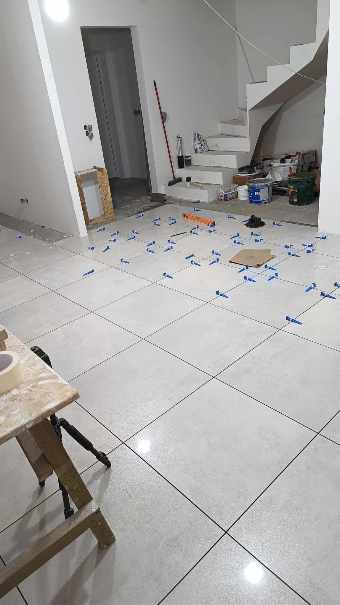 Jasne, kwadratowe płytki podłogowe w trakcie układania, z niebieskimi krzyżykami dystansowymi. W tle widoczne schody i narzędzia glazurnicze.