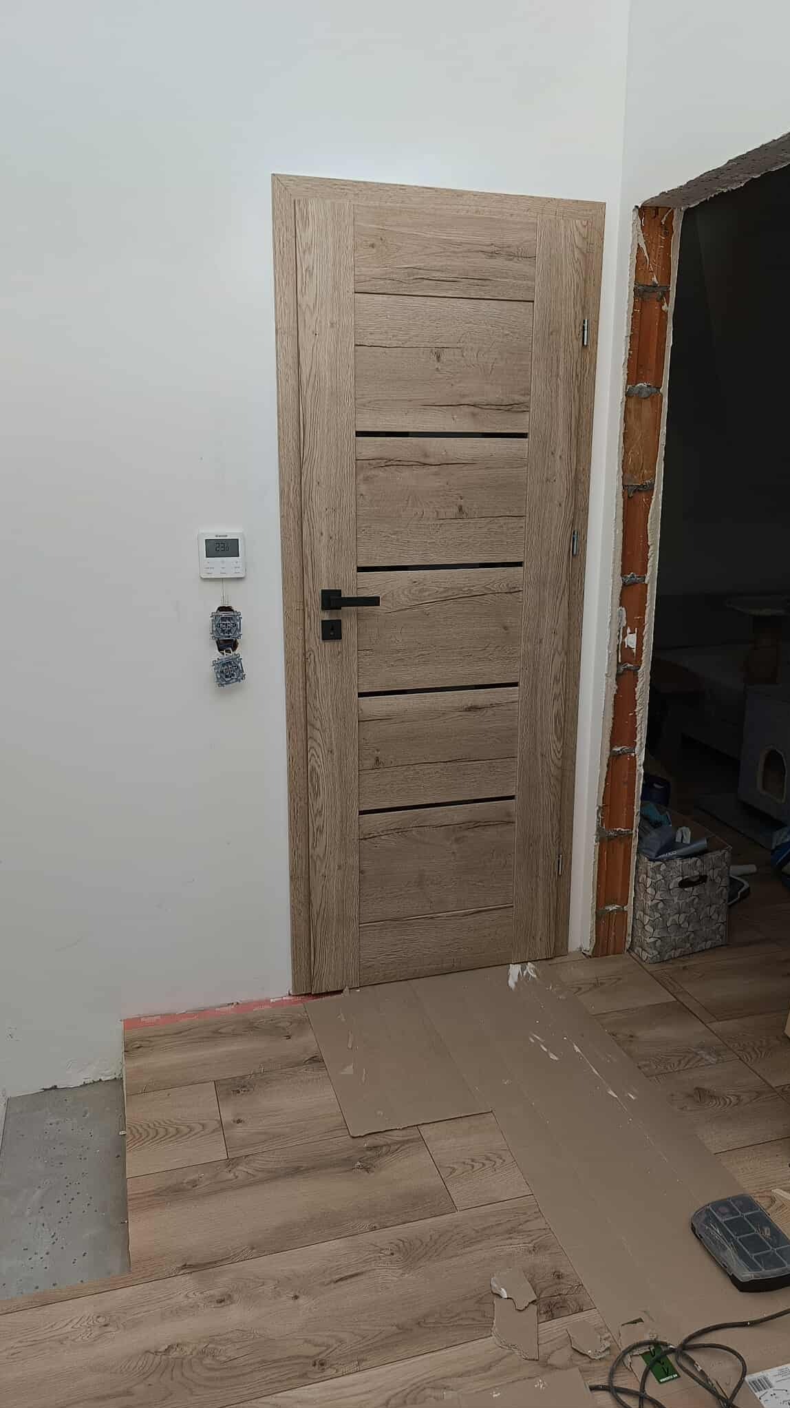 Montaż drzwi wewnętrznych z drewnianą okleiną i czarną klamką, widoczne niedokończone prace wykończeniowe podłogi i ściany, gniazdka elektryczne bez obudowy.