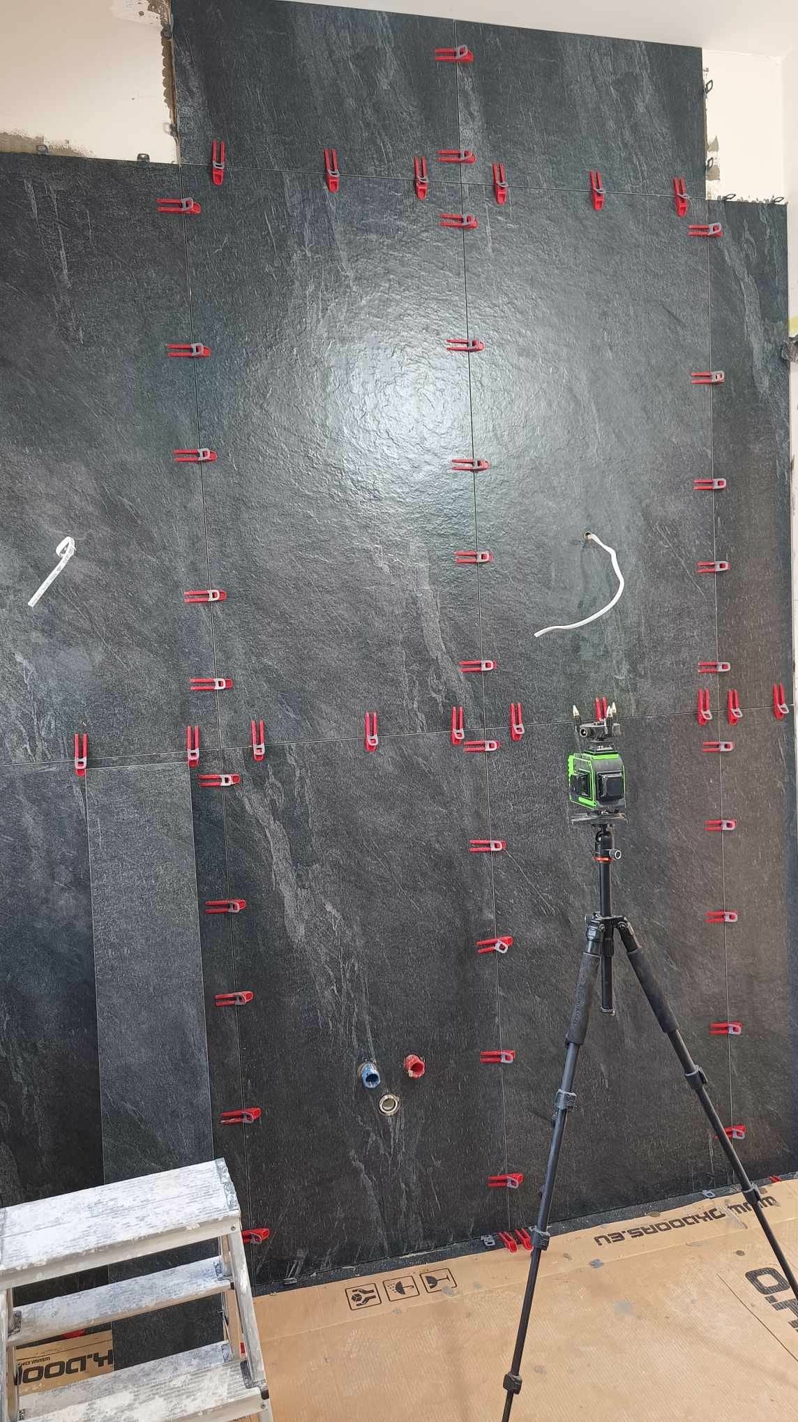 Układanie ciemnych płytek na ścianie z użyciem poziomicy laserowej i klipsów poziomujących. Widoczny fragment drabiny i karton na podłodze.