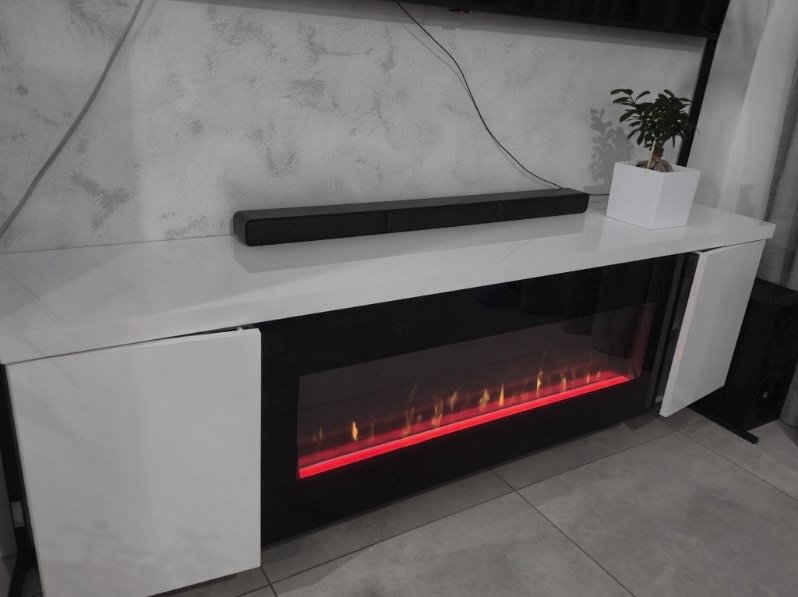 Biała szafka RTV z elektrycznym kominkiem, na której stoi soundbar i roślina w doniczce. Ściana w tle z efektem betonu. Nowoczesny design.