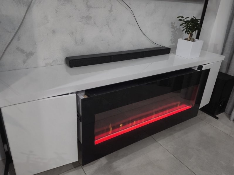 Biała szafka RTV z elektrycznym kominkiem i soundbarem na tle szarej ściany. Widoczny fragment rośliny w białej doniczce. Nowoczesny design.