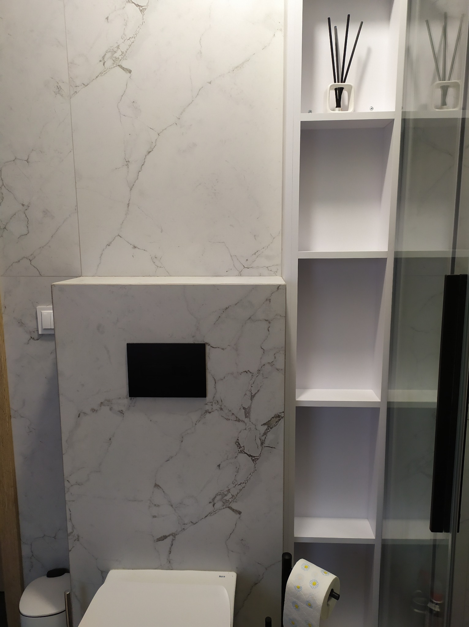 Nowoczesna łazienka z marmurowymi płytkami, podwieszaną toaletą i białą zabudową meblową z półkami, na których stoją dekoracyjne dyfuzory zapachowe.