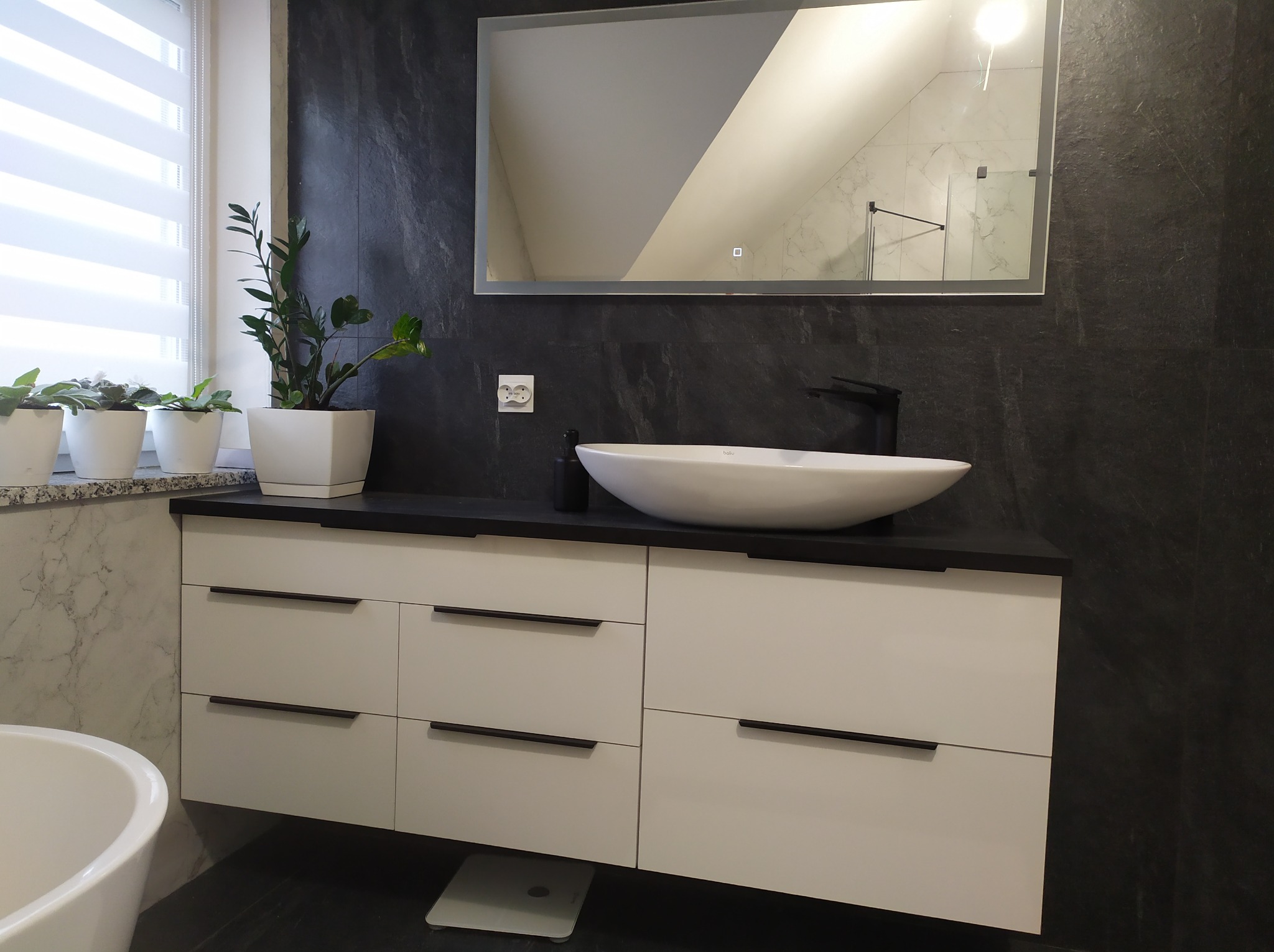 Nowoczesna łazienka z białą szafką pod umywalkę, czarnym blatem i armaturą. Na blacie owalna umywalka i roślina. W tle lustro z podświetleniem i ciemna ściana.