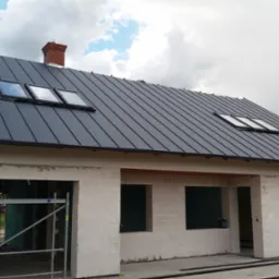 Nowy, grafitowy dach z blachy na surowym budynku z oknami dachowymi i ceglanym kominem. Widoczne rusztowanie i elementy konstrukcyjne.