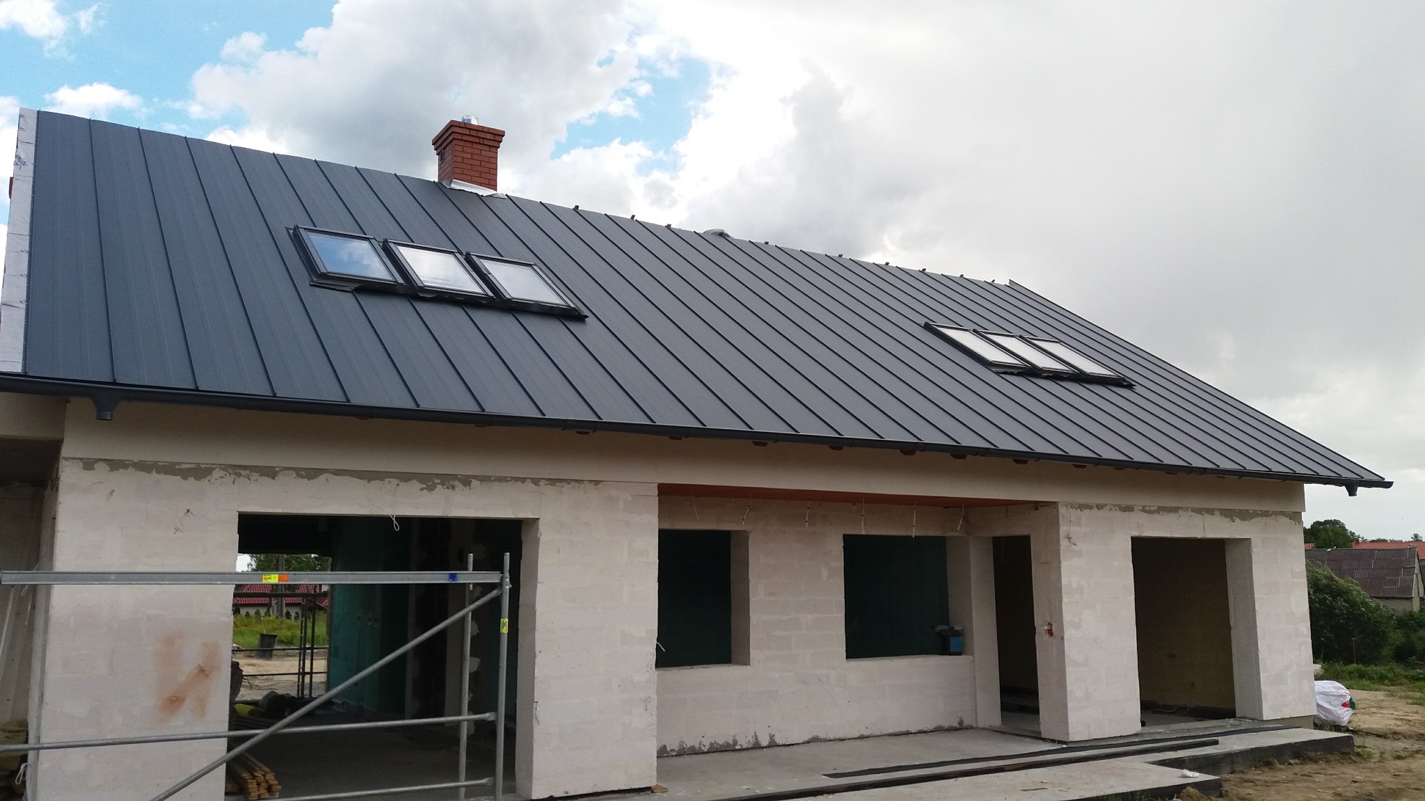 Nowy, grafitowy dach z blachy na surowym budynku z oknami dachowymi i ceglanym kominem. Widoczne rusztowanie i elementy konstrukcyjne.