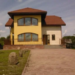 Żółto-biały dom z brązowym dachem i ceglanym podjazdem, ozdobiony kamieniami na trawniku.