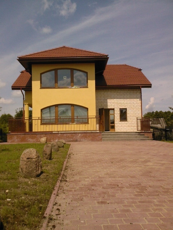 Żółto-biały dom z brązowym dachem i ceglanym podjazdem, ozdobiony kamieniami na trawniku.