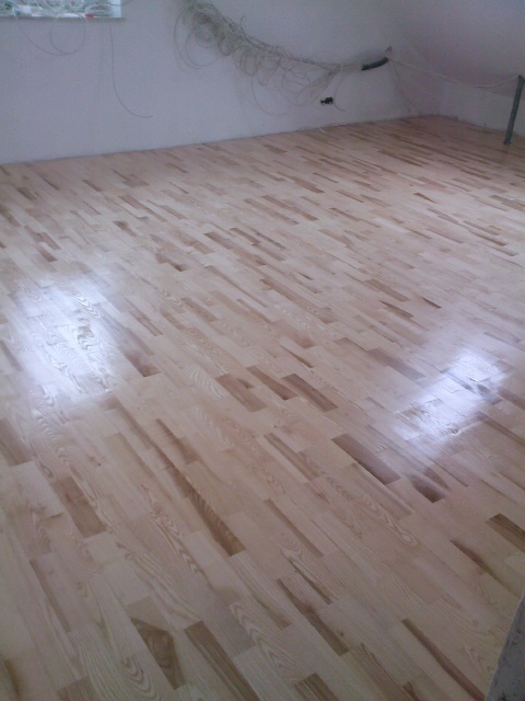 Jasna, drewniana podłoga parkietowa ułożona pod kątem w nowo wykończonym wnętrzu poddasza, widoczne kable elektryczne zwisające z sufitu.