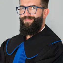 Portret brodatego mężczyzny w todze adwokackiej i okularach, z zegarkiem na nadgarstku, uśmiechającego się do kamery na neutralnym tle.