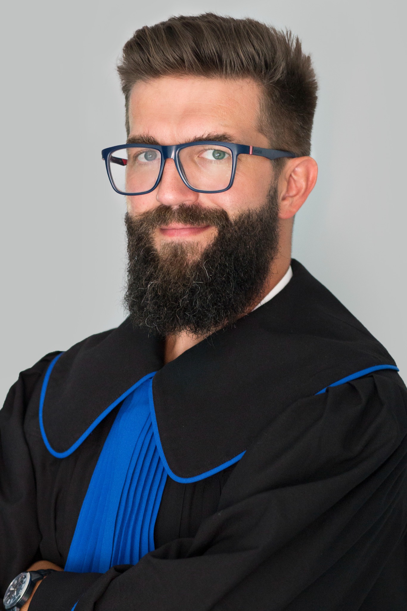 Portret brodatego mężczyzny w todze adwokackiej i okularach, z zegarkiem na nadgarstku, uśmiechającego się do kamery na neutralnym tle.