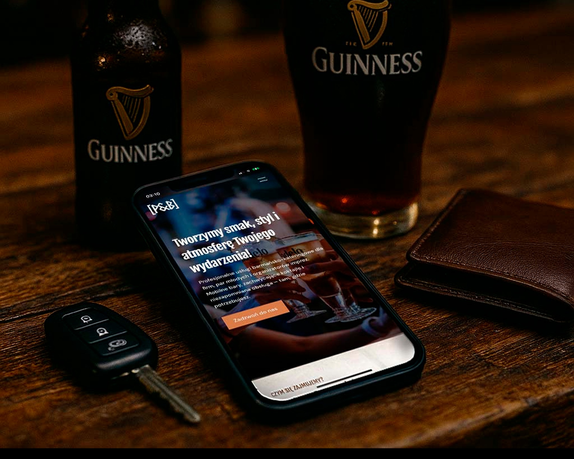 Smartfon wyświetla stronę z ofertą barmańską na tle Guinessa, kluczyka samochodowego i portfela na drewnianym stole. Aranżacja sugeruje profesjonalizm i luksus.