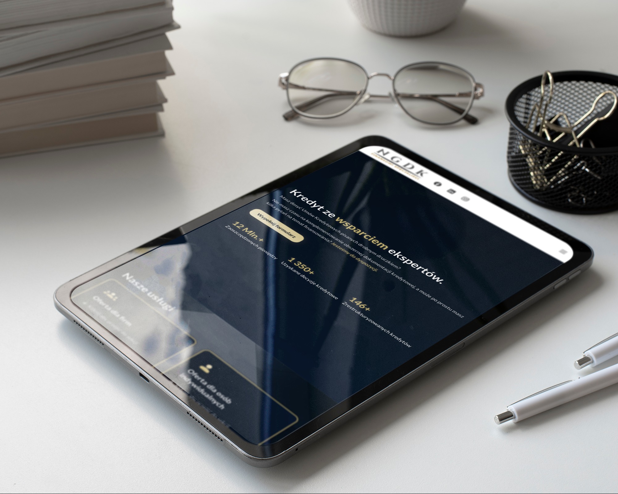 Tablet prezentuje stronę internetową firmy N.G.D.K. z ofertą 'Kredyt ze wsparciem ekspertów'. Widoczne statystyki: 12M+, 1350+, 146+.