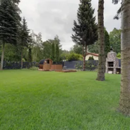 Zadbany ogród z równo przystrzyżonym trawnikiem, drewnianą sauną beczką, jacuzzi i murowanym grillem. W tle wysokie drzewa i czarne ogrodzenie.