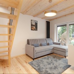 JARO-INVESTMENT - Jasne wnętrze pokoju z drewnianymi schodami, szarą sofą i ozdobną lampą. Widoczne drewniane wykończenie sufitu i podłogi, okno z widokiem na zewnątrz.