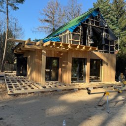 JARO-INVESTMENT - Budowa domu szkieletowego w trakcie: drewniana konstrukcja, okna wstawione, widoczne membrany dachowe i podkonstrukcja tarasu. Na pierwszym planie piasek i butelka wody.