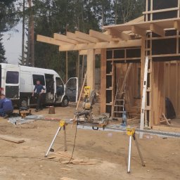 JARO-INVESTMENT - Budowa domu szkieletowego w lesie: widok na konstrukcję z drewna, narzędzia pracy (piła) i ekipę montażową przy białym busie. Etap wznoszenia ścian i zadaszenia.