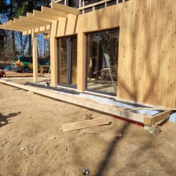 JARO-INVESTMENT - Budowa domu szkieletowego: montaż drewnianej elewacji i pergoli. Widoczne okna, fundament i pracujący robotnicy. Słoneczny dzień, naturalne oświetlenie.