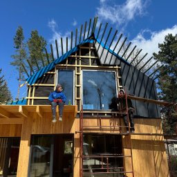 JARO-INVESTMENT - Budowa domu: dwaj robotnicy przy montażu drewnianej elewacji i dachu z membraną. Jeden siedzi na belce, drugi na rusztowaniu. Błękitne niebo w tle.