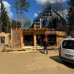 JARO-INVESTMENT - Budowa domu modułowego: drewniana konstrukcja, duże okna, robotnicy na rusztowaniu montują dach pokryty niebieską folią. Na pierwszym planie osoba w czerwonej bluzie.