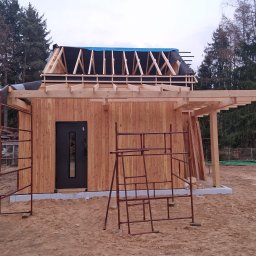 JARO-INVESTMENT - Budowa domu szkieletowego w trakcie: widoczne drewniane ściany, konstrukcja dachu z membraną, rusztowania. Dom z ciemnymi drzwiami, otoczony drzewami.