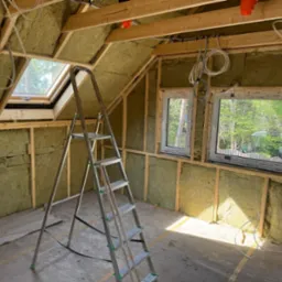 Poddasze z oknami połaciowymi i ścianami wypełnionymi izolacją z wełny mineralnej. Widoczna drabina, drewniana konstrukcja i instalacje elektryczne.