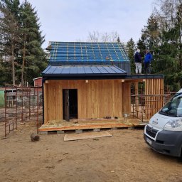 JARO-INVESTMENT - Budowa domu modułowego z drewnianą elewacją i niebieskim dachem. Dwóch pracowników na dachu, widoczny samochód firmowy i rusztowania.