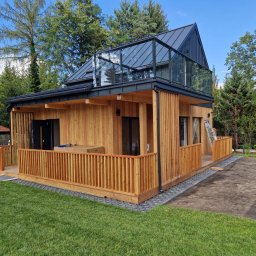 JARO-INVESTMENT - Nowoczesny dom modułowy z drewna z przeszkloną nadbudówką i drewnianym tarasem, otoczony zielenią. Widoczne detale wykończenia i drabina oparta o ścianę.