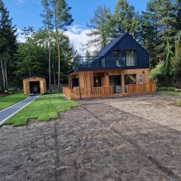 JARO-INVESTMENT - Nowoczesny dom modułowy z drewnianą elewacją i czarnym dachem w otoczeniu drzew. Widoczny fragment trawnika w trakcie układania i mała drewniana szopa.
