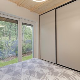 JARO-INVESTMENT - Nowoczesna sypialnia: duże okno z widokiem na zieleń, przesuwana szafa z czarnymi obramowaniami, drewniany sufit i geometryczna pościel w odcieniach szarości.